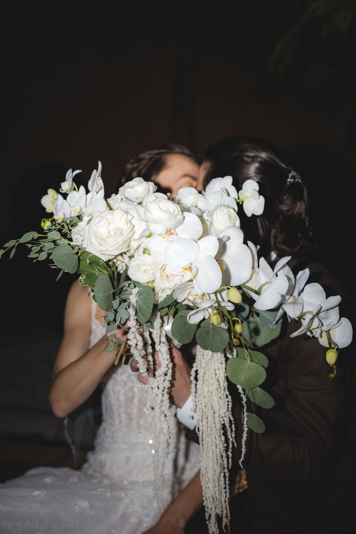 A stunning white bridal bouquet featuring orchids, roses and cascading amaranthus against a dark background.