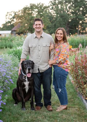 A couple stands with their large black and white dog in a blooming garden at sunset.