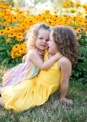 Two children in yellow and pink outfits laying in grass near bright orange flowers on a sunny summer day.