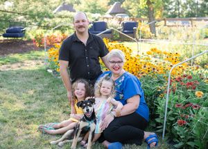 A family sits together with their dog in a garden filled with bright yellow and orange flowers on a sunny day.