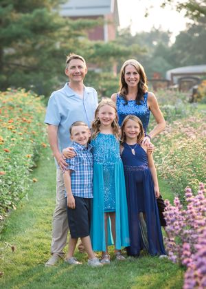 A family poses together in coordinating blue outfits among blooming wildflowers in a garden setting at sunset.