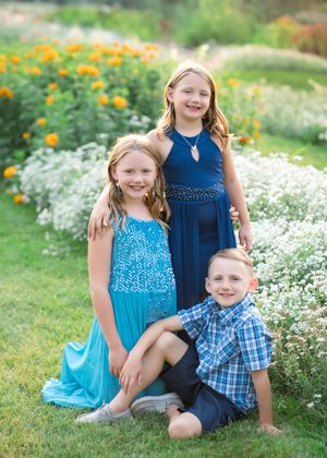 Children sit together in a garden with white and orange flowers wearing blue and plaid outfits on a summer day.