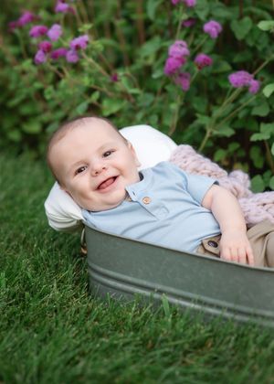 Baby lies in a metal tub outdoors surrounded by purple flowers and green foliage in the garden.