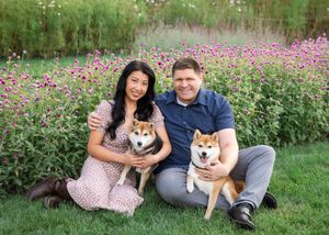 A couple and their Shiba Inu dog pose together in a field of pink cosmos flowers on a sunny day.