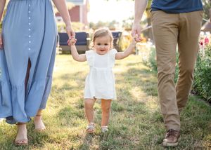 A toddler in a white dress walks while holding hands with parents in a sunny garden setting.