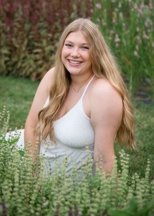 A person in a white top poses outdoors among flowering plants and greenery in a natural garden setting.