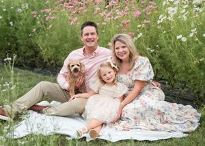 A family sits together with their small dog on a white blanket surrounded by blooming cosmos flowers.