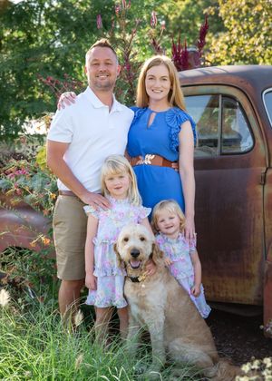 Family poses for outdoor portrait session with their golden doodle dog next to vintage rustic truck in summer setting.