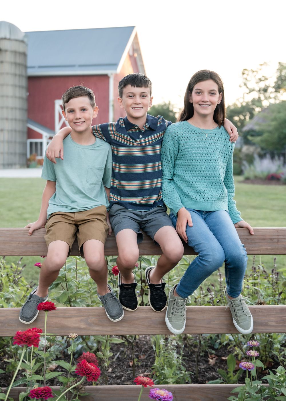 Three children sit together on a wooden bench in front of a garden with red flowers and a red barn in the background.