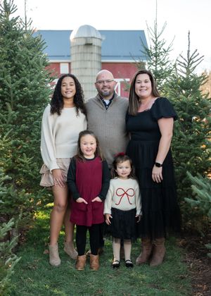 A family poses together between evergreen trees with a red barn and silo in the background during a holiday photo session.