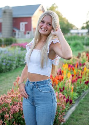 A woman in a white crop top and jeans poses in front of colorful flower gardens near a red barn.