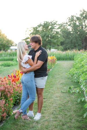 A couple embraces in a garden path surrounded by vibrant red snapdragon flowers and lush greenery on a summer day.