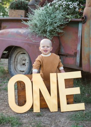 A series of gold glittery letters spelling ONE arranged outdoors against a rustic red truck and greenery backdrop.