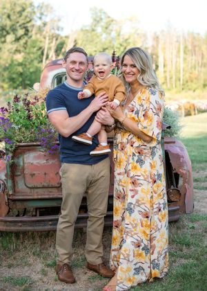 A family poses together in front of a rustic red truck with flowers, wearing casual attire in an outdoor garden setting.