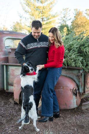 A couple and their black and white border collie dog pose together near potted evergreen trees in winter.