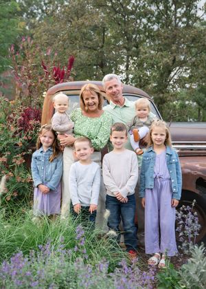 A group of children stand together in front of a vintage truck with purple wildflowers blooming in the foreground.