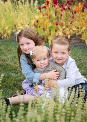 Three children sit together on grass with colorful autumn foliage in the background.