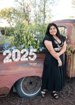 A graduate poses in a black dress next to vintage truck wheels with '2025' signage in an outdoor setting.