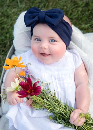 Baby in white dress and navy bow headband holds colorful wildflowers while lying on a blanket outdoors.