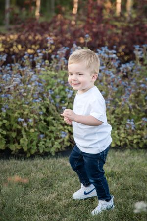 A small child in a white shirt and jeans plays outdoors near flowering purple plants in a garden setting.
