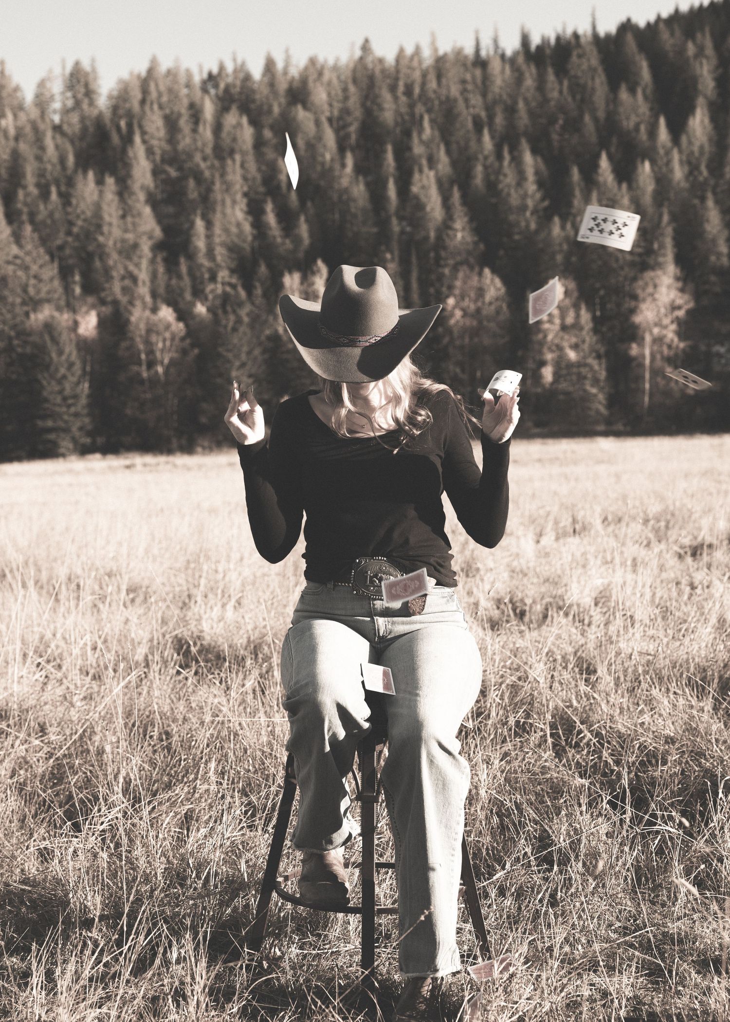 Woman in cowboy hat sitting on stool tossing playing cards in a sepia-toned outdoor field.