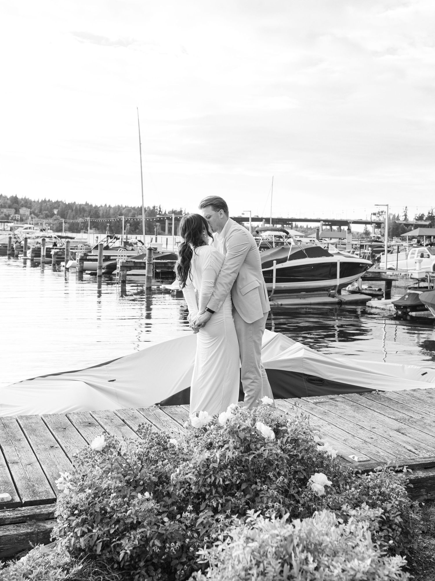 Black and white photograph of a couple embracing on a wooden dock overlooking a marina filled with boats.
