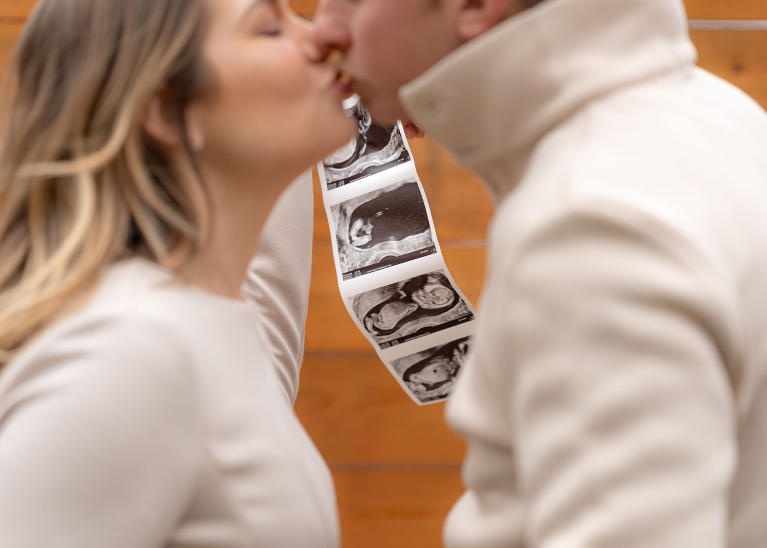 A couple in cream colored sweaters share an intimate moment against a wooden wall.