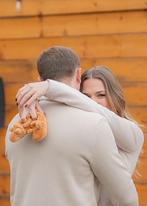 A couple in cream sweaters embraces while holding something in front of wooden walls.