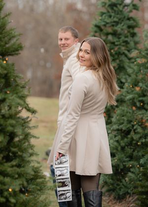 A couple shares a joyful moment while holding photos in front of evergreen trees.
