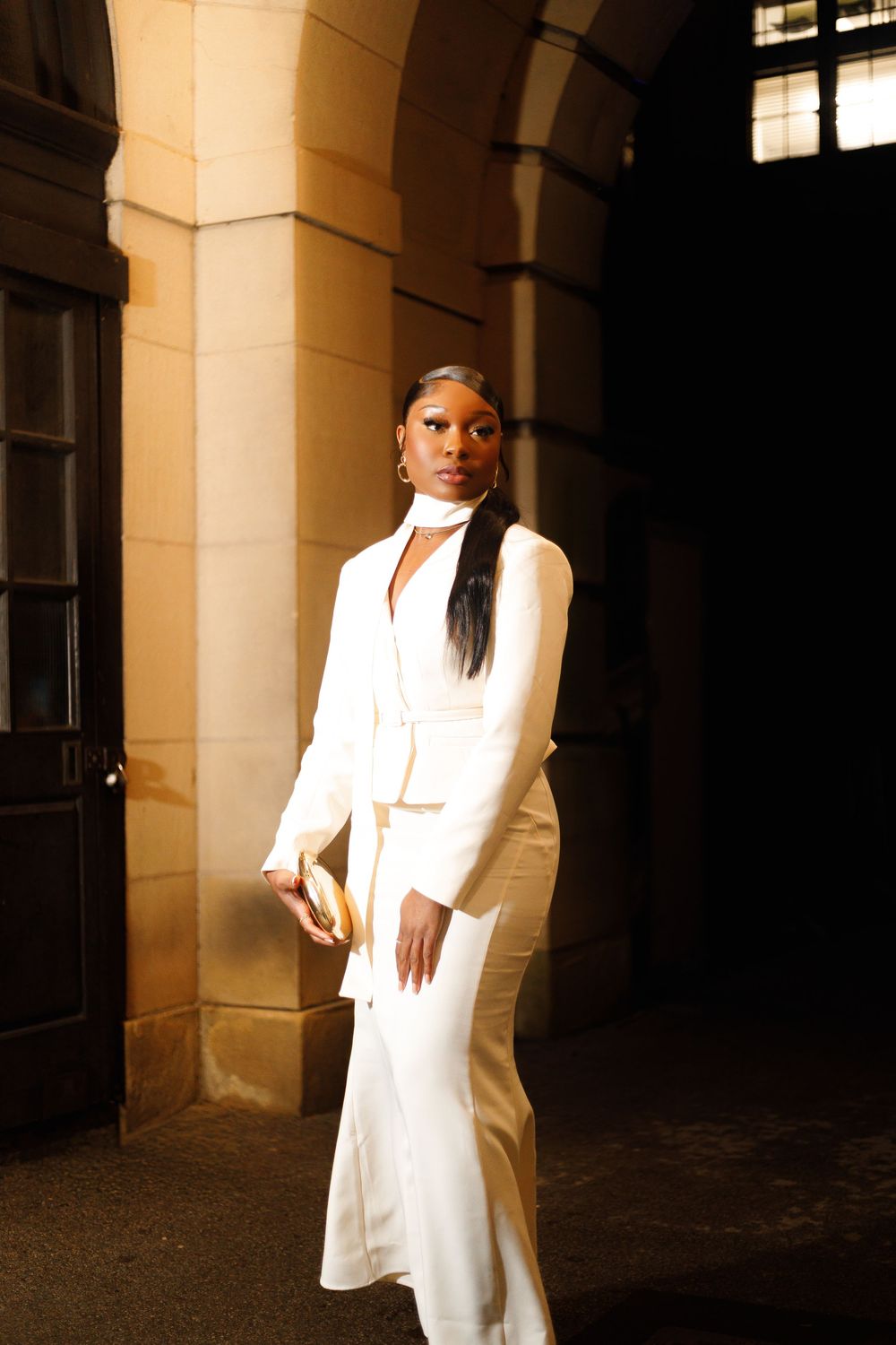 A person in a flowing white outfit poses dramatically in a hallway with stone columns and arches.