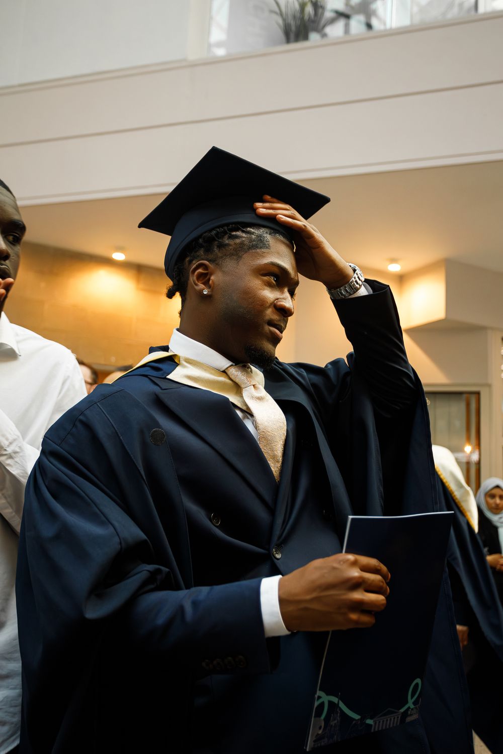 Graduates in black caps and gowns stand together during a graduation ceremony in a warmly lit indoor space.