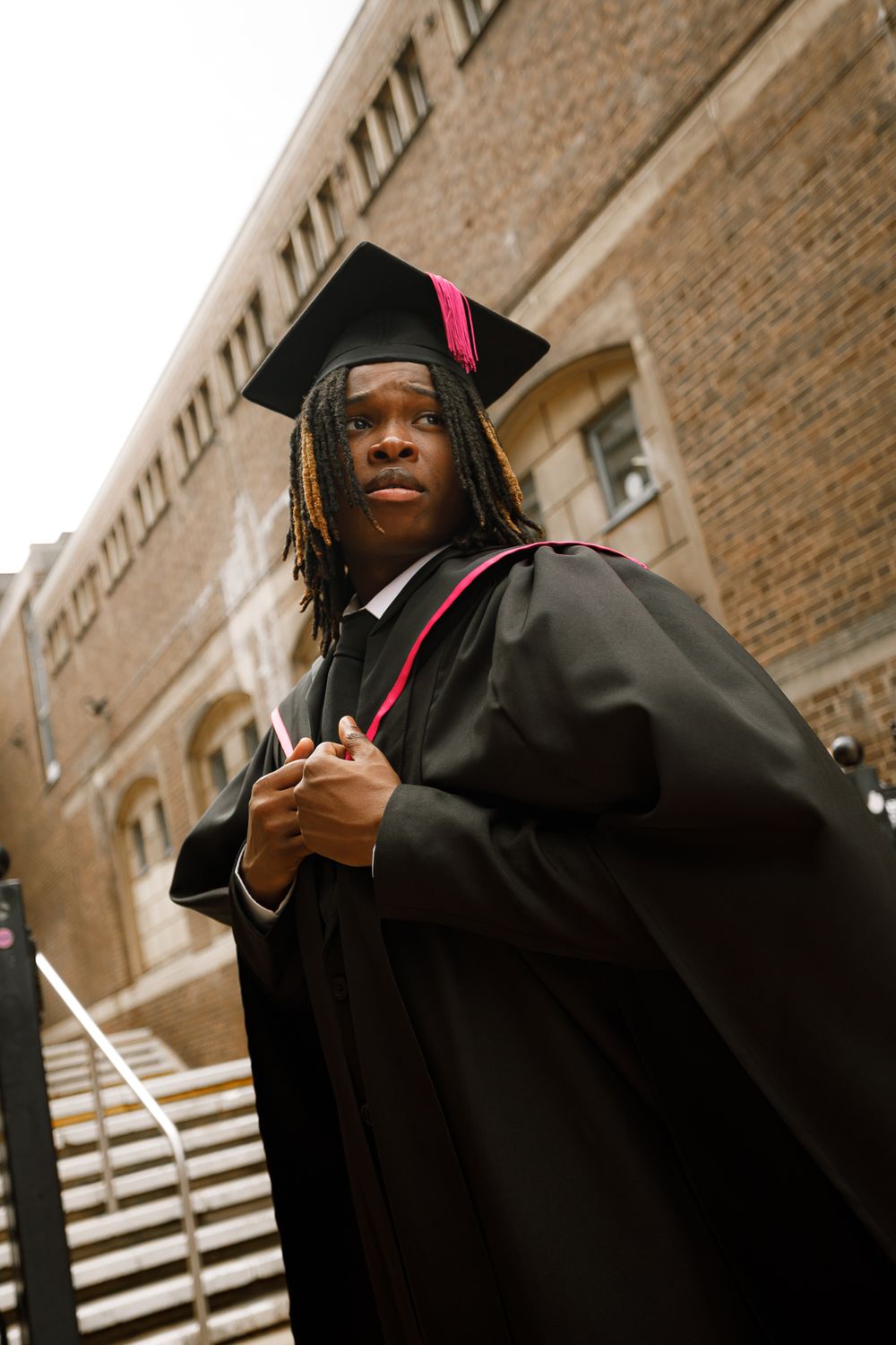 Graduate in black cap and gown holding diploma against brick building exterior on graduation day.