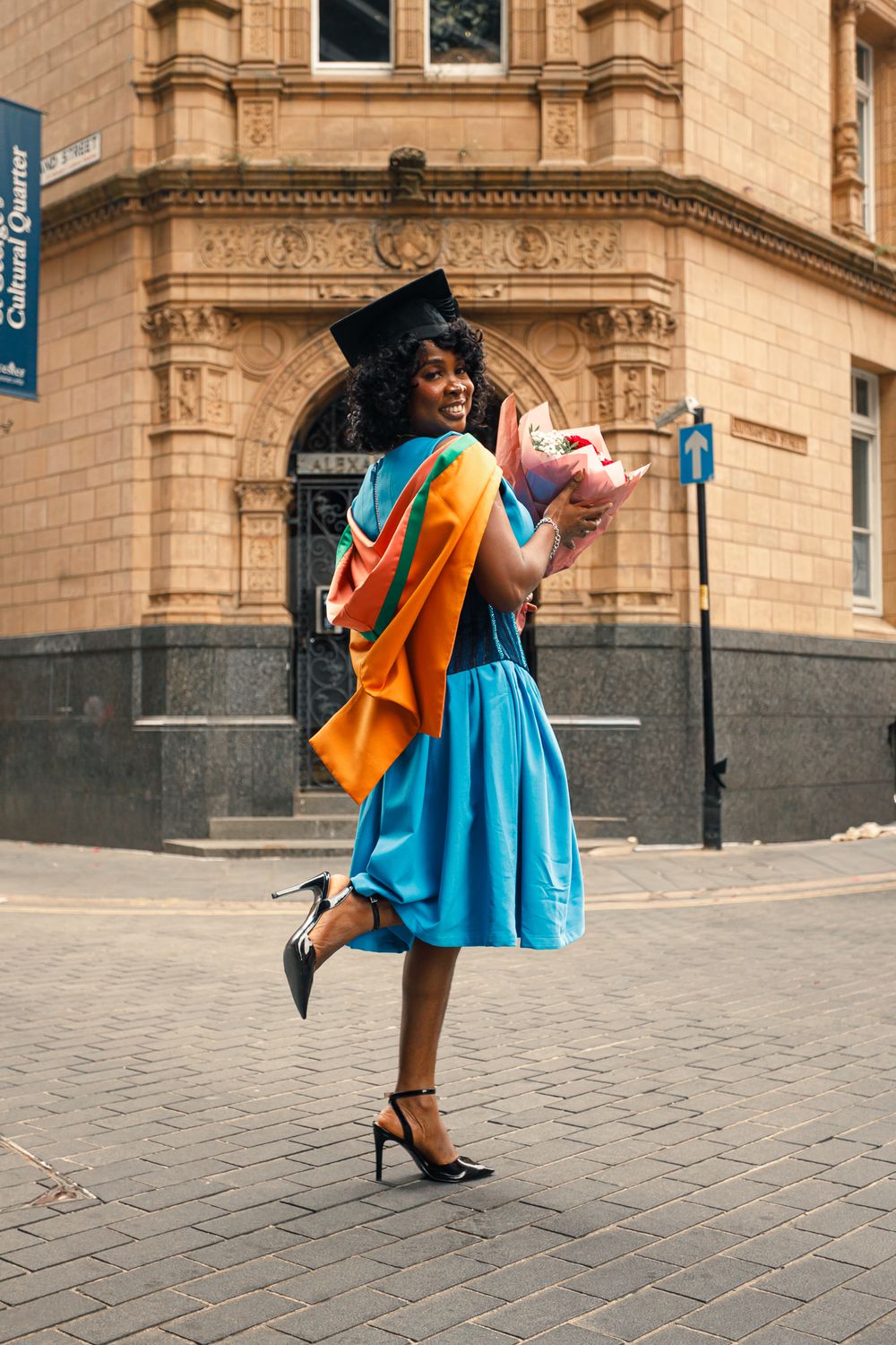 Two friends in bright orange and blue dresses embrace and dance joyfully on a cobblestone street near historic buildings.