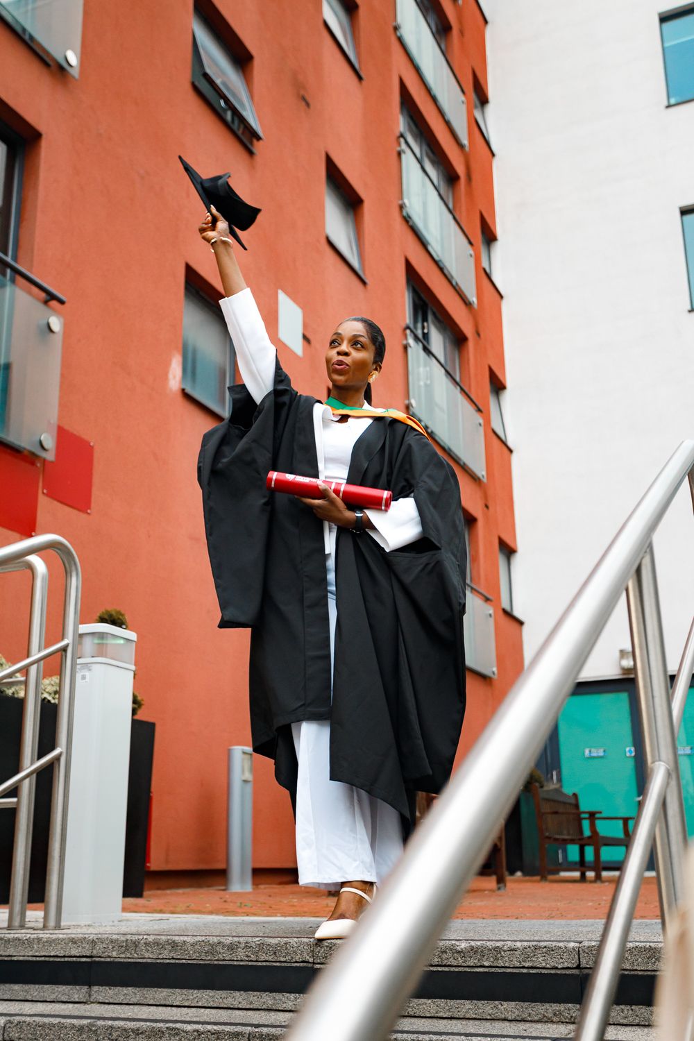 Graduate in black academic gown celebrates on steps of orange building while holding diploma and raising cap in air.