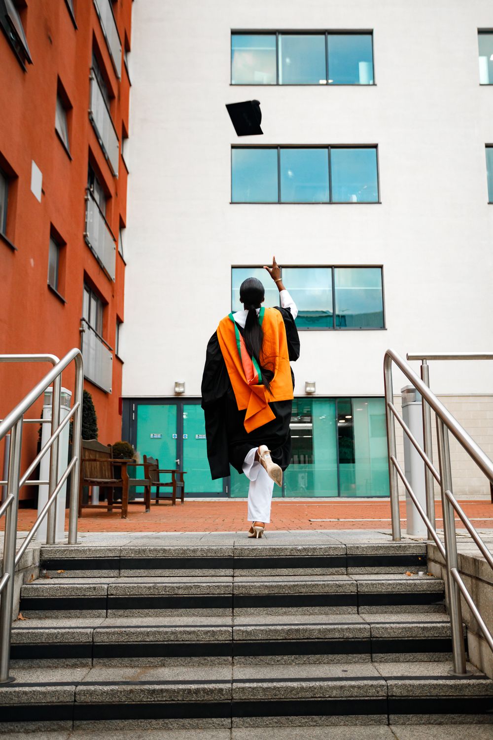 A person in an orange safety vest throws a black hat while sitting on concrete steps outside a modern building.