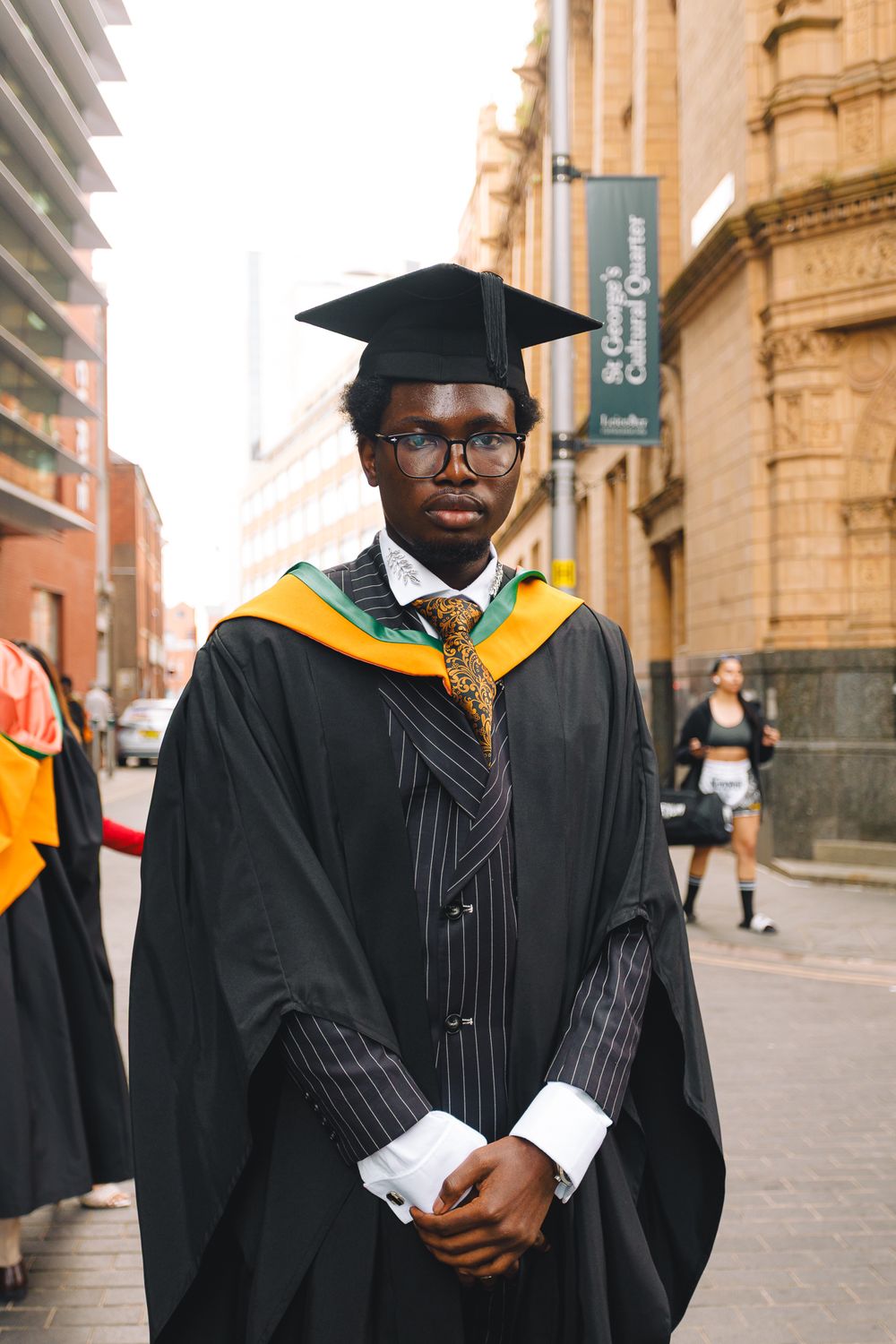 Graduate in black academic regalia and gold stole walking on a city street during commencement celebrations.