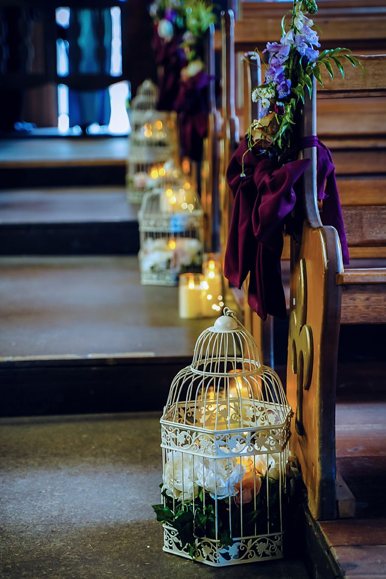 Decorative white birdcages with flowers and fairy lights line a church aisle for a romantic wedding ceremony.