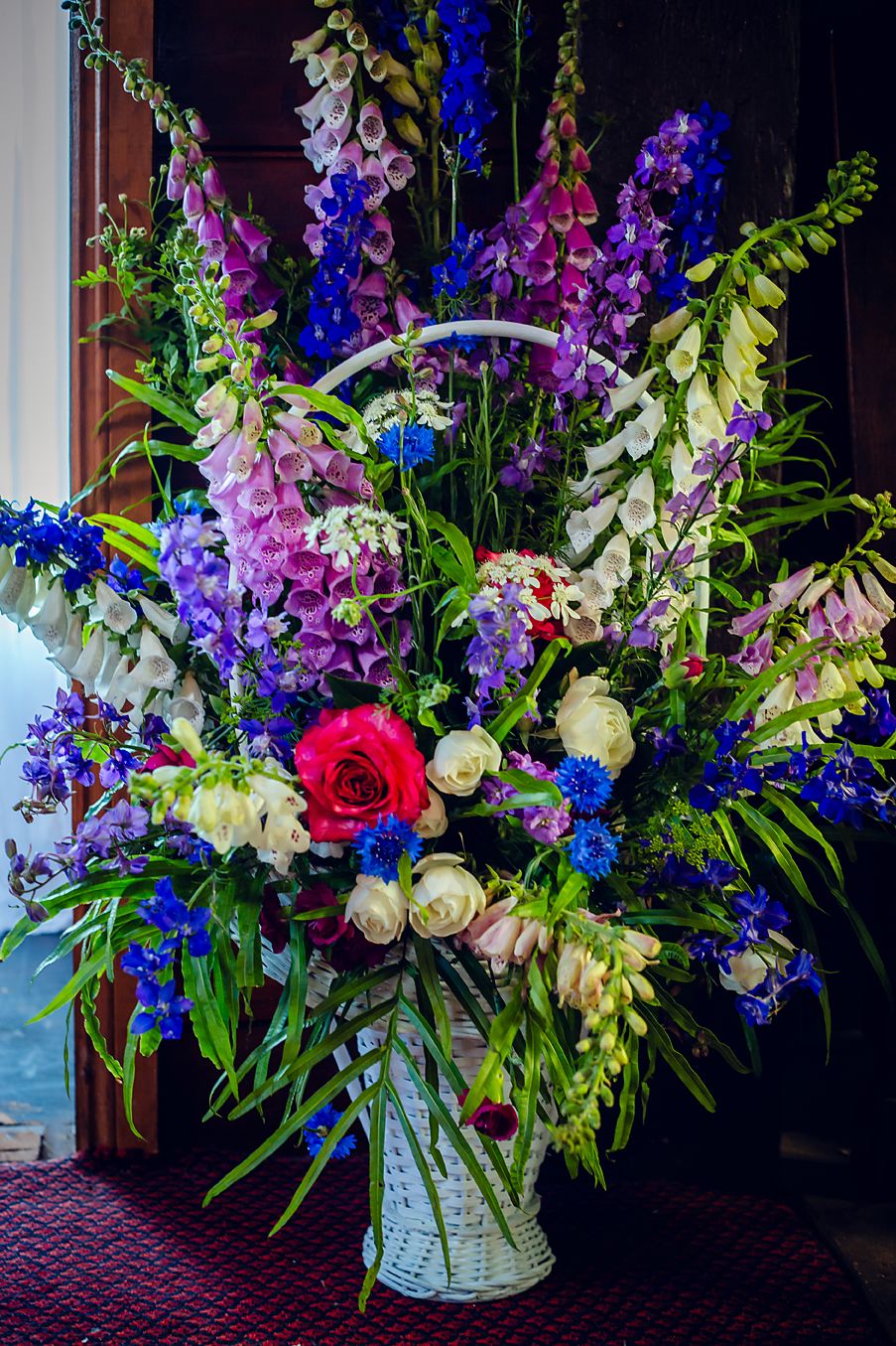 Vibrant floral arrangement with purple delphinium, red roses, and white blooms in an elegant vase against a dark background.