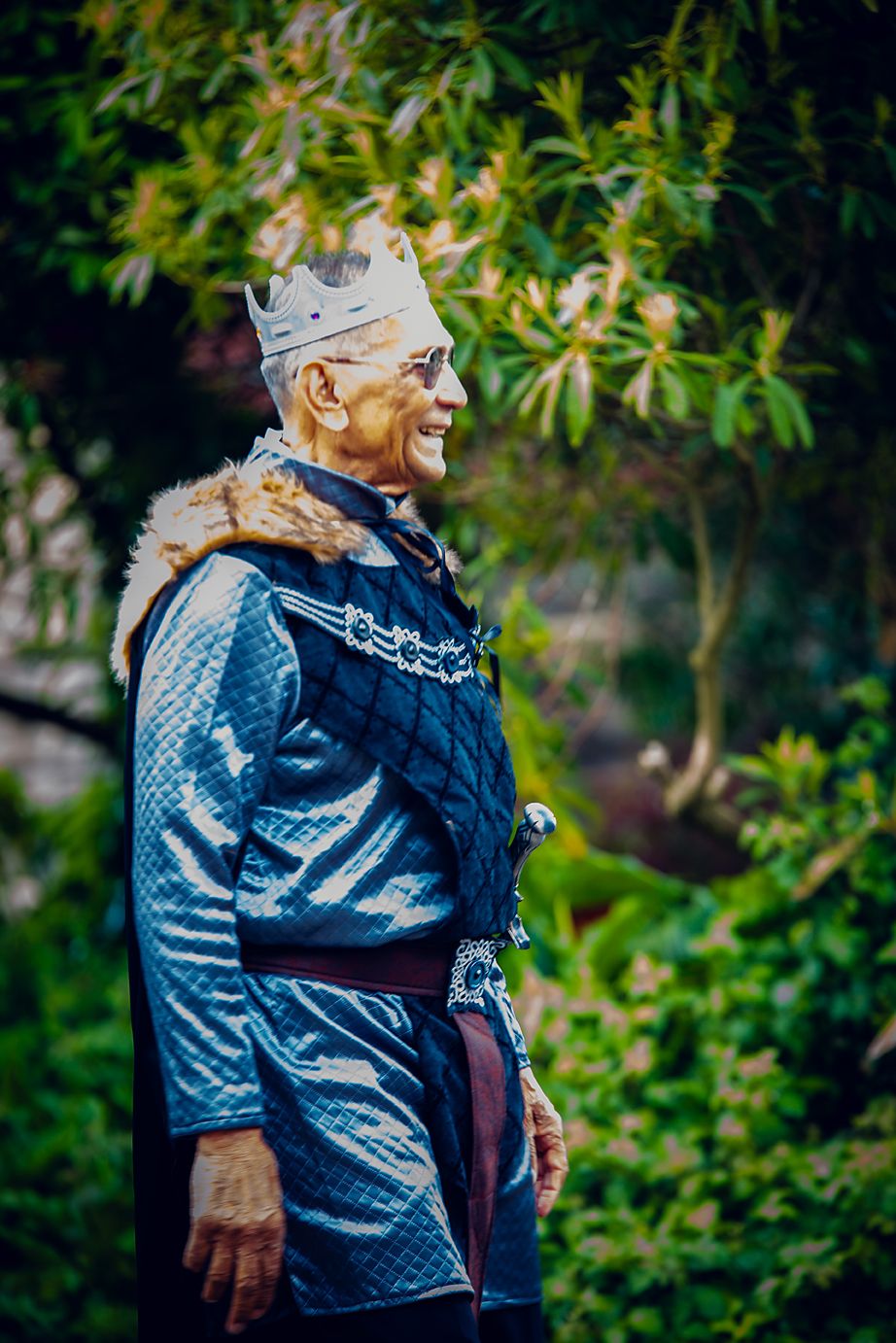 A person wearing a blue quilted jacket and glasses smiles while standing in front of green foliage outdoors.