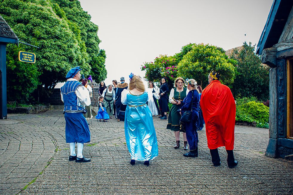 A group of people in colorful costumes gather on a paved walkway surrounded by green trees and bushes.