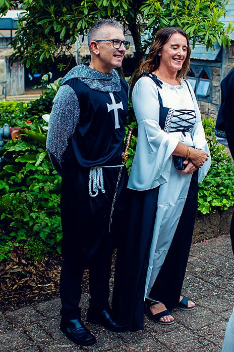 Two people in black and white outfits laugh together while standing in front of green foliage outdoors.