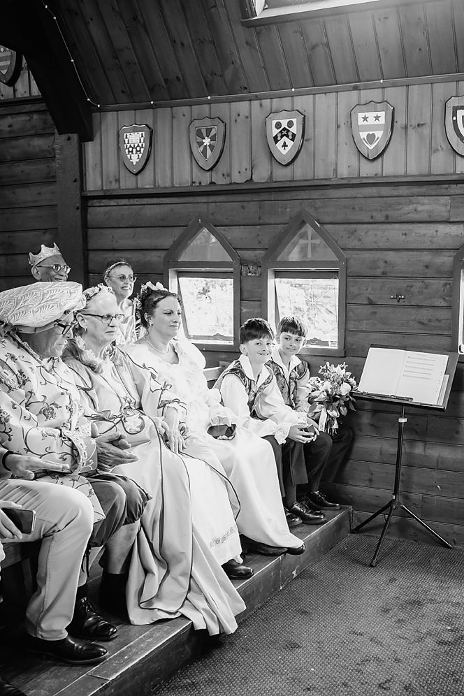 Black and white photograph showing people seated on wooden benches in a rustic cabin interior with shields on the wall.