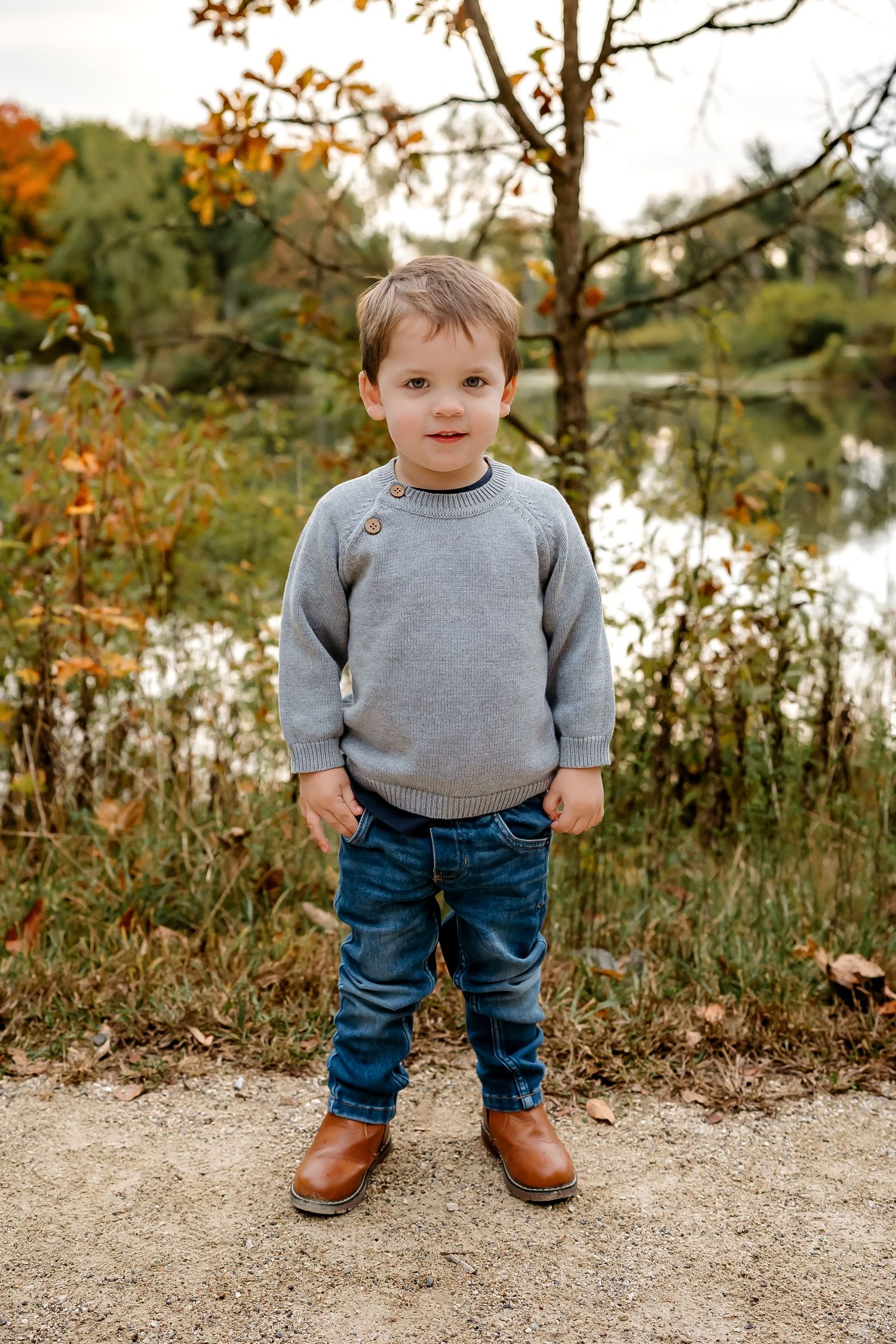 Small child wearing gray sweater and jeans stands on path during autumn with water and fall foliage in background.