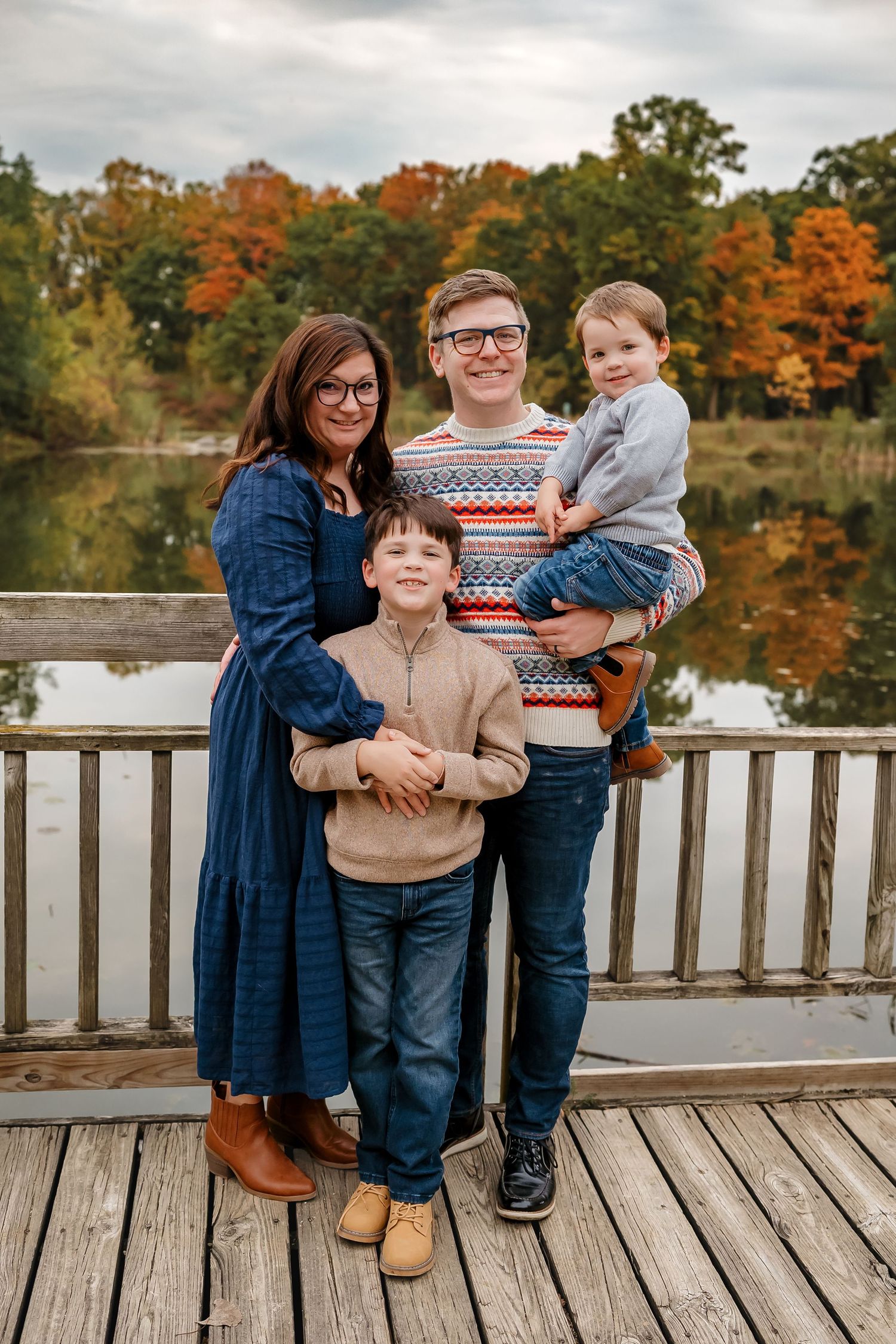 A family stands together on a wooden dock with vibrant fall foliage and a calm lake reflecting the autumn colors behind them.