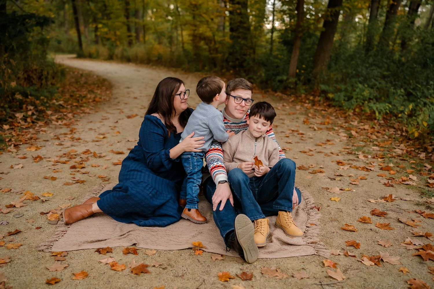 Family sits together on leaf-covered path in autumn forest during outdoor photo session.