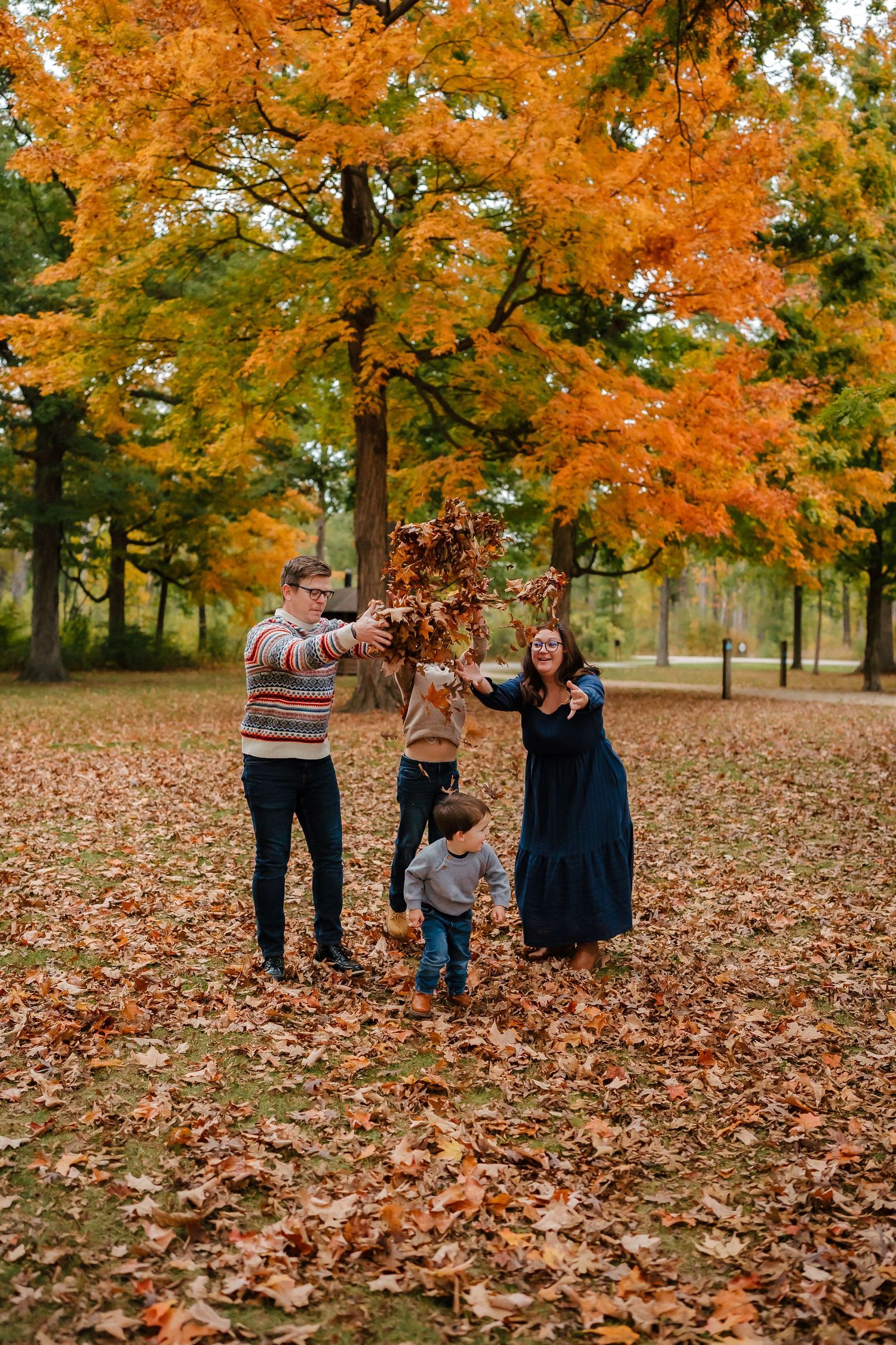 A family plays together in a park covered with vibrant orange autumn leaves under maple trees.