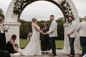 A wedding ceremony takes place under a white decorative arch outdoors with guests gathered around.
