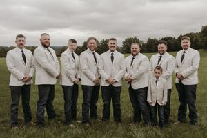 Groomsmen in light colored jackets and dark pants pose together in a line on a grassy field.