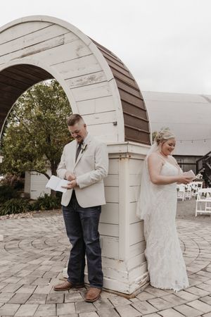 A couple stands on opposite sides of a white archway during their wedding ceremony at an outdoor venue.