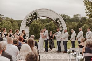 A panoramic view of an outdoor wedding ceremony with guests seated in white chairs under a decorative arch.